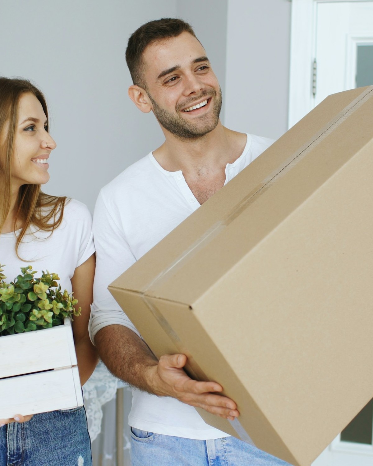 Two people smiling as they carry a box and a plant crate into a new home